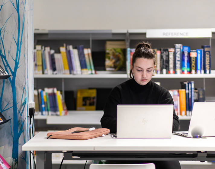 Student sitting at a desk with two laptops in the library at Glasgow Kelvin College’s Easterhouse campus, focused on studying in front of a reference bookshelf. Student sitting at a desk with two laptops in the library at Glasgow Kelvin College’s Easterhouse campus, focused on studying in front of a reference bookshelf.